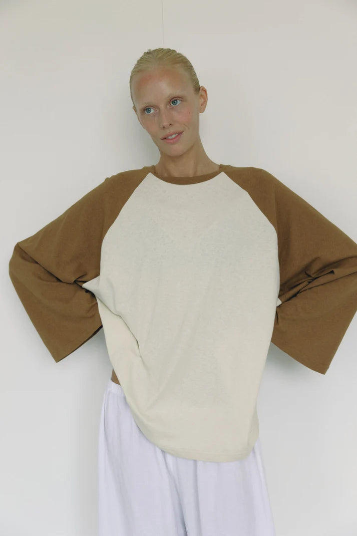 A model wearing a long-sleeve raglan shirt with a cream-colored body and brown sleeves, paired with white pants, standing against a plain off-white wall.